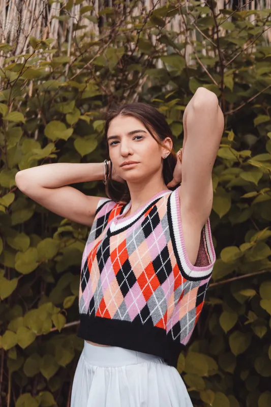 Argyle knit vest, arms raised, direct gaze in soft light