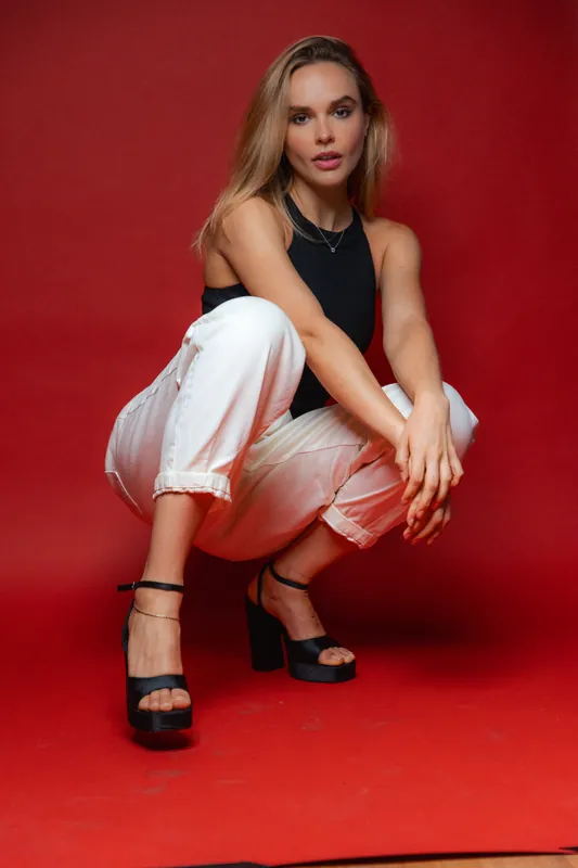 Black bodysuit and white trousers, crouching on a red studio backdrop