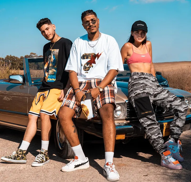 Three models in streetwear posed against a vintage convertible in an open Israeli field