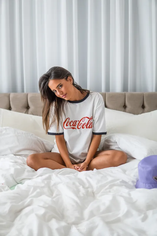 Oversized Coca-Cola tee, sitting cross-legged on a white bed, Tel Aviv